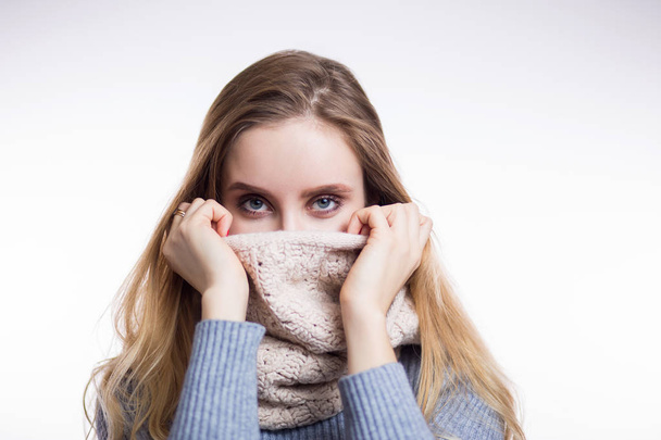 Winter portrait of beautiful blonde woman in sweater and scarf on a white background. Girl covers her face with snood and looking at the camera - Foto, afbeelding