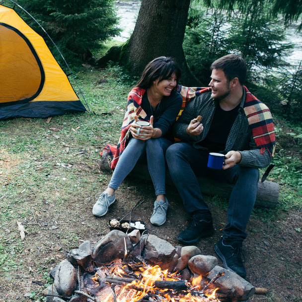 Pärchen sitzen am Lagerfeuer, trinken Tee und erzählen Geschichten. Zelt und Geländewagen im Hintergrund. Vereinigung mit der Natur - Foto, Bild