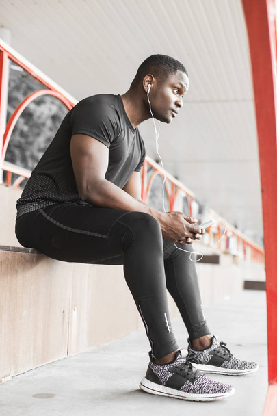 Young male jogger athlete training and doing workout outdoors in city. a black man resting after a workout and listening to music and watching a sports watch - Photo, image