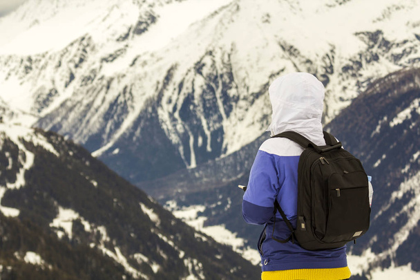 ΠΙΣΩ ΟΨΗ της τουριστικής πεζοπόρος σε ζεστά ρούχα με σακίδιο που στέκεται πάνω σε ψηλά βουνά, βραχώδη καλυμμένα με χιόνι και θυελλώδη συννεφιασμένο ουρανό αντίγραφο χώρου φόντο. - Φωτογραφία, εικόνα