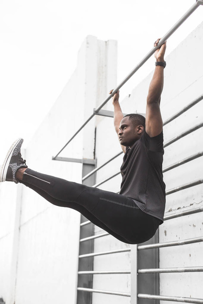 Concentrated african american sportsman warming up and stretching legs on pier, pulls up on a horizontal bar - 写真・画像
