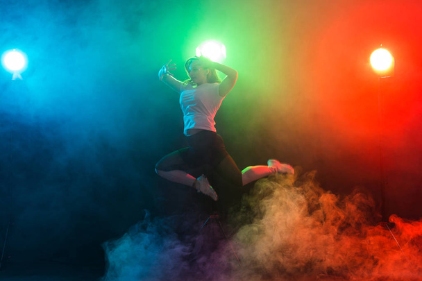 Dansen, sport, jazz funk en mensen concept - jonge vrouw sprong in de duisternis onder kleurrijk licht - Foto, afbeelding