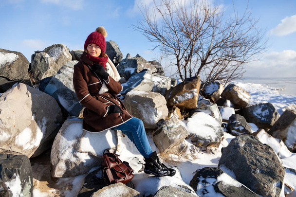 Foto caliente de invierno. Retrato de una hermosa modelo joven en sombrero de punto y ropa de abrigo oliendo y sentado en piedra en el polo norte, detrás de montañas cubiertas de nieve. alegre humor alegre
. - Foto, Imagen