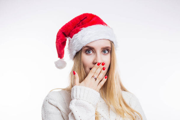 Portrait of surprised attractive young woman in red Santa hat and beige sweater on white background. A girl with a bright manicure closes her mouth with her hand and opens her eyes - 写真・画像