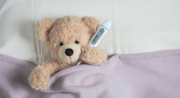 Fever, cold, flue concept. Lindo peluche en la cama, cubierto con una manta caliente, sosteniendo un termómetro
 - Foto, imagen