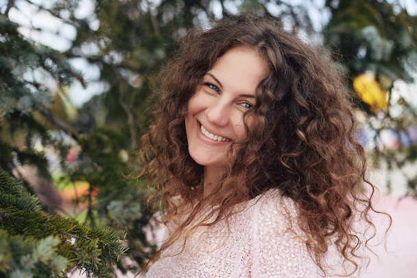 Nyári portré fiatal kaukázusi göndör hajú nő, mosolygós boldog fényképezőgép tökéletes fogak és a kék szemek, fákkal körülvett. Szép nő pózol a szabadban. Életmód, boldogság koncepciója. - Fotó, kép