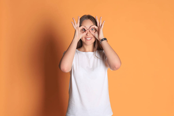 Young woman in basic clothing smiling looking at camera through holes made with fingers isolated over yellow background. - Foto, afbeelding