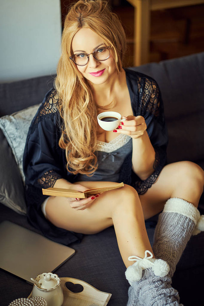 молода красива блондинка сидить на сірому дивані і читає книгу
 - Фото, зображення