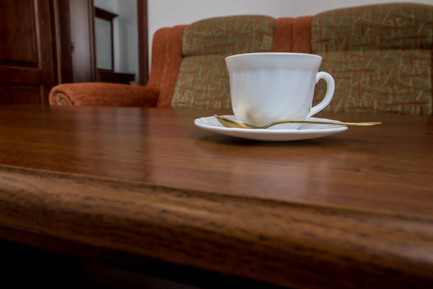 белая кружка с блюдцем и ложкой стоит на деревянном столе рядом с диваном
 - Фото, изображение