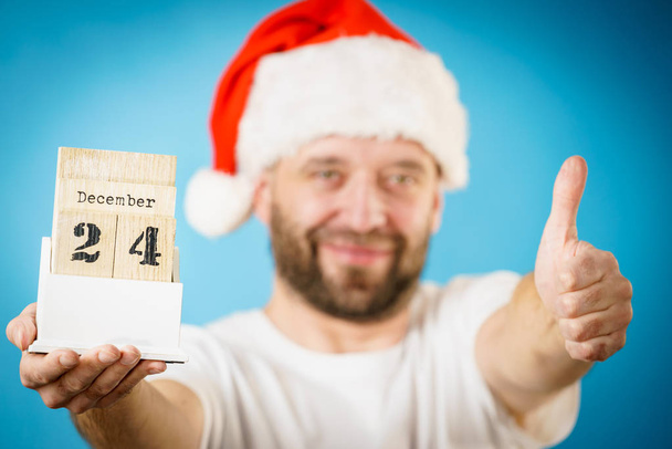 Man wearing santa claus christmas hat holding calendar with 24 december date, on blue - Photo, image