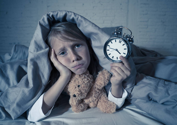Cute sleepless little girl lying in bed showing alarm clock looking tired having sleeping troubles staying asleep at night or waking too early in Children Insomnia Anxiety and Sleep Disorders. - Foto, imagen