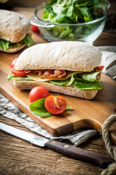 Ciabatta sandwich with arugula salad, bacon and yellow cheese on wooden board. - 写真・画像