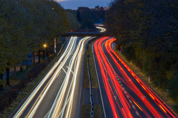 lange Belichtungsspuren des Verkehrs auf einer Autobahn im Abendlicht, die durch eine bewaldete Landschaft fahren, von einer Brücke aus betrachtet - Foto, Bild