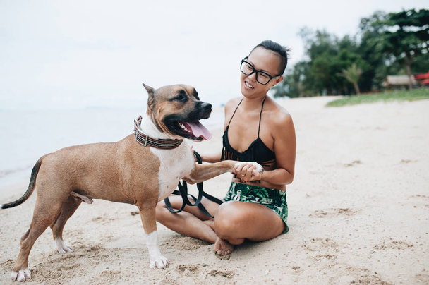 Cheerful pretty young woman in eyeglases sitting and hugging her dog on the beach - Foto, imagen