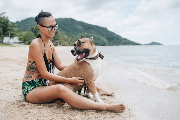 Wesoły dość młoda kobieta w eyeglases siedzi i przytulanie jej pies na plaży - Zdjęcie, obraz