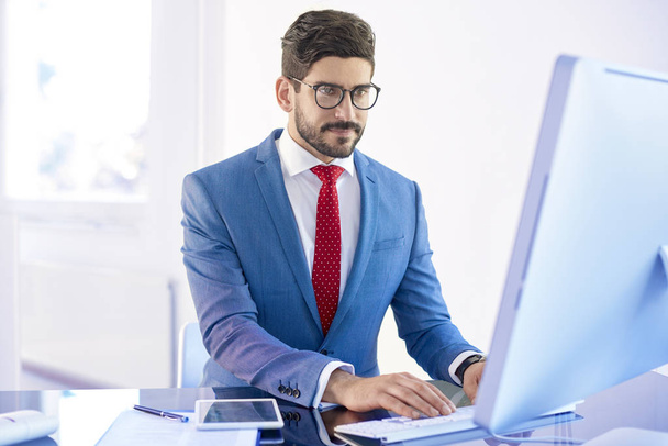 Portrait of young businessman wearing suit while sitting in front of computer and working on investment project.  - Фото, зображення