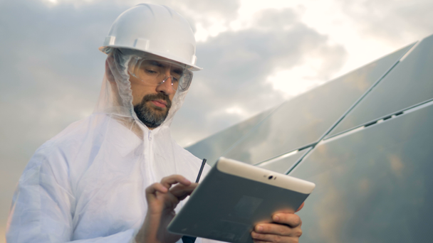 Man in protection wear is operating a tablet next to a massive solar installation - Imágenes, Vídeo