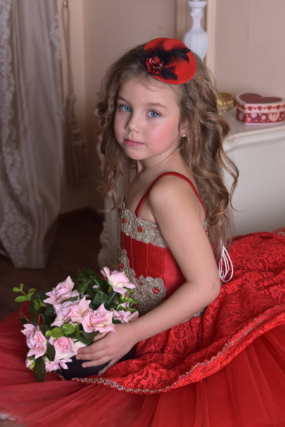 portrait of a little girl princess in a red dress and a hat sits on the sofa with a box of roses - Φωτογραφία, εικόνα