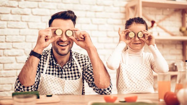 Cook Food at Home. Father Feeds Daughter. Happy Family. Father's Day. Girl and Man Cook Food. Spend Time Together. Sliced Tomatoes on Board. Man with Vegetables. Sliced Cucumbers near Eyes. - 写真・画像