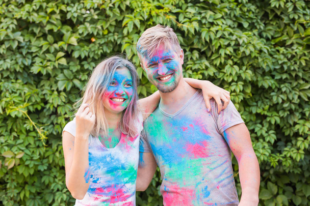 Holiday and people concept - Smiling woman and man posing with multicolored powder on their faces - Photo, image