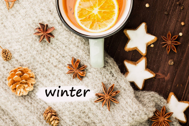 Θέμα "Χειμώνας". Ζεστό τσάι με μπαχαρικά, πορτοκάλι, γλυκάνισο, μπισκότα σε σχήμα αστέρι, το πιπέρι και το γκρι κασκόλ σε ξύλινο υπόβαθρο. Επίπεδη lay, θέα από ψηλά με το χειμώνα κείμενο σε λευκό. - Φωτογραφία, εικόνα