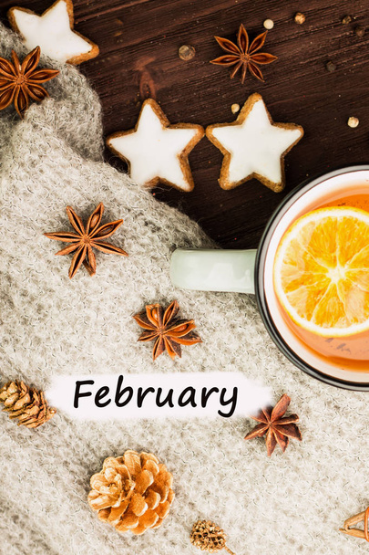Θέμα "Χειμώνας". Ζεστό τσάι με μπαχαρικά, πορτοκάλι, γλυκάνισο, μπισκότα σε σχήμα αστέρι, το πιπέρι και το γκρι κασκόλ σε ξύλινο υπόβαθρο. Επίπεδη lay, θέα από ψηλά με κείμενο Φεβρουαρίου σε λευκό. - Φωτογραφία, εικόνα