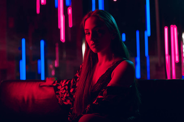 Sensual young woman sitting on a leather sofa lit by red neon light and with a wall of neon tubes in a background - Foto, imagen