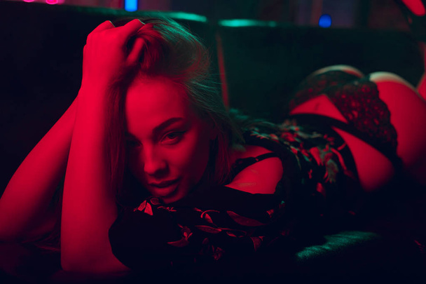 Attractive young woman lying bended out in neon lights with alluring look into the camera - Фото, изображение