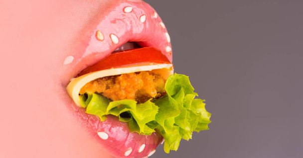 Fast food. Kobiece usta i burger wewnątrz. Zamknąć usta różowe kobieta z pomidor, ser, kotlet, sałata wewnątrz. Uwodzicielski warg kobiece z sezamem pełne sałatka. Usta pyszne - Zdjęcie, obraz