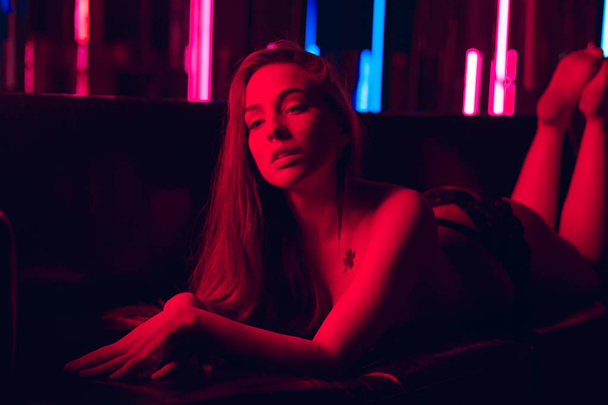 Piękna młoda kobieta, oświetlone przez czerwone światło neon sexy, pochylając się nad leżąc nago lub nago na kanapie obok ściany z rur neonowych - Zdjęcie, obraz