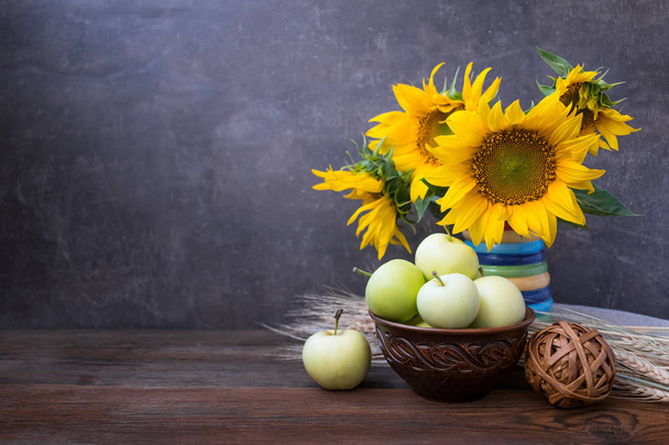 Autumn harvest, natural products, Greeting card concept. Still life with beautiful sunflower flower, green apples in clay brown bowl, wheat spikelets on wooden rustic background. - 写真・画像