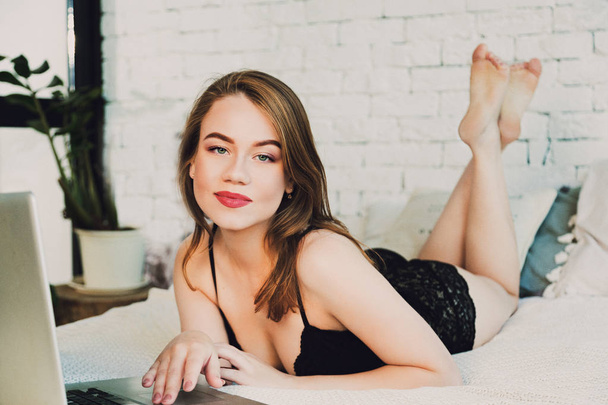 Portrait of a charming sexy woman in lingerie lying on the bed playing social network or social media on notebook or laptop online - Фото, изображение