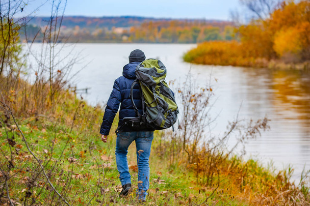 A tourist with a backpack walks next to the river in autumn. A beautiful landskape of nature in yellow-orange colours. - Foto, Imagen