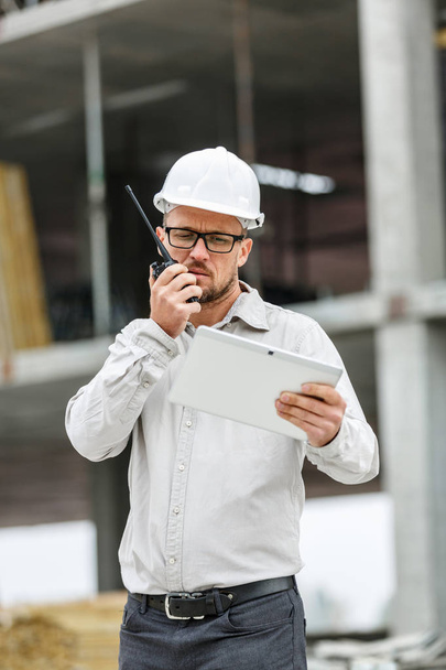 Male head engineer wearing white safety hardhat with walkie talkie and tablet inspecting construction site. Technology, development and construction industry concept - Foto, Imagen
