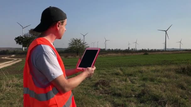 Technical engineer in safety vest work on tablet near working wind turbine towers on windy sunny day. Technician using touch-sensitive tablet in fields. Worker on power farm near wind mill power plant - Footage, Video