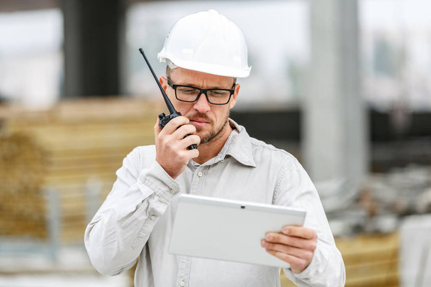 Male head engineer wearing white safety hardhat with walkie talkie and tablet inspecting construction site. Technology, development and construction industry concept - Фото, изображение