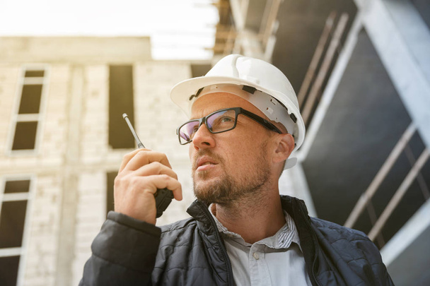 Male head engineer wearing white safety hardhat using walkie talkie while inspecting construction site. Development and construction industry concept - Foto, immagini