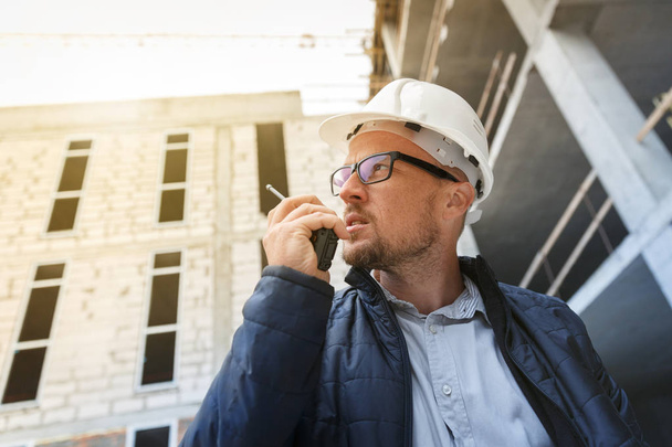 Male head engineer wearing white safety hardhat using walkie talkie while inspecting construction site. Development and construction industry concept - Photo, Image