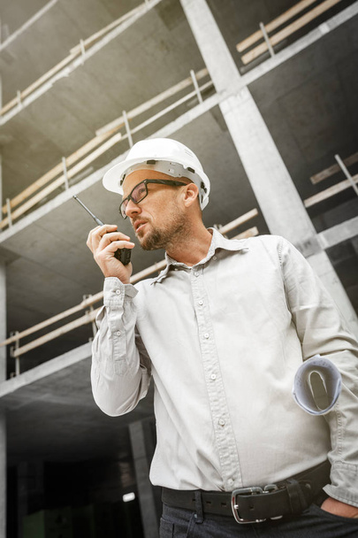 Male head engineer wearing white safety hardhat with walkie talkie and blueprint inspecting construction site. Development and construction industry concept - Photo, image