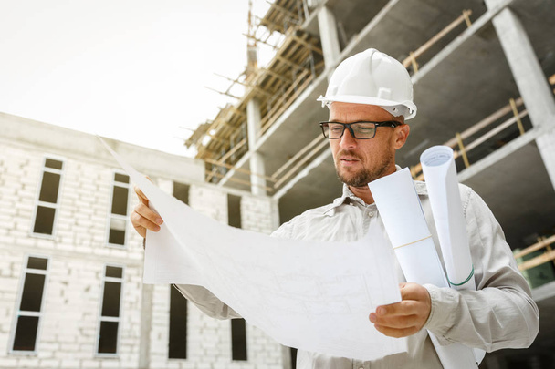 Construction engineer in a white hardhat inspecting blueprints on a construction site. Development and construction industry concept - Foto, Bild