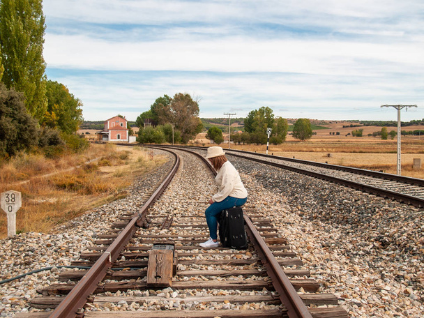 Eine traurige blonde Frau mit Strohhut auf den Bahngleisen sitzt im Herbst auf einem Koffer - Foto, Bild