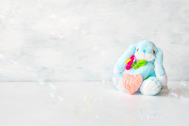 Cute toy soft blue bunny with a flower and pink thread heart with soap bubbles on light background. Birthday card, Valentine's Day, mother's day, Easter background. - Zdjęcie, obraz