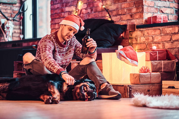 Ένα κομψό άνδρα κρατώντας ένα μπουκάλι μπύρας ενώ κάθεται με το χαριτωμένο σκυλί σε ένα διακοσμημένο καθιστικό Χριστούγεννα. - Φωτογραφία, εικόνα