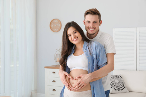 Pregnant woman and her husband showing heart with hands at home - Фото, изображение