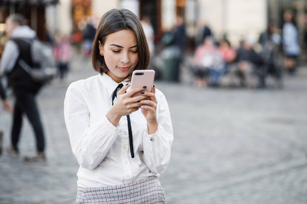 Portrait of a beautiful woman using mobile phone while standing on blurred street background. Girls using an application in her smart phone. - Фото, изображение