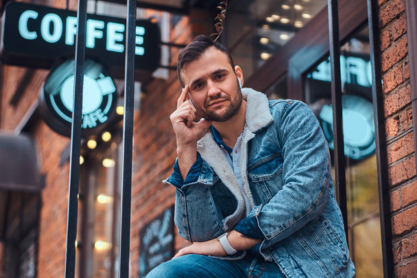 Μια συλλογισμένος κομψό άνδρα που φοράει ένα σακάκι τζιν με ασύρματα ακουστικά κρατώντας takeaway καφέ έξω από το μαγαζί. - Φωτογραφία, εικόνα