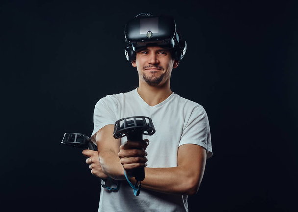 Retrato de un hombre con cerdas vestido con camisa blanca y gafas de realidad virtual y sostiene joysticks, mirando a la cámara
. - Foto, imagen