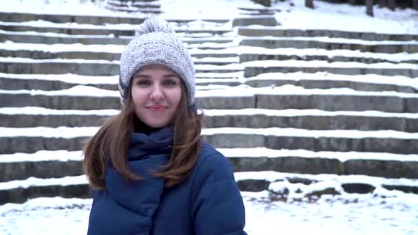 Όμορφη γυναίκα στο χειμερινό καπέλο χαμόγελα στέκεται έξω από το χιόνι στο δάσος με φόντο χιονισμένο σκάλες. Πορτρέτο του ένα όμορφο κορίτσι ψάχνει στην κάμερα. Χειμώνα πορτρέτο ενός νεαρού κοριτσιού. Το - Πλάνα, βίντεο