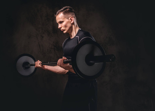 Focused young sportsman holding a barbell and doing exercise on biceps. Isolated on dark background. - Photo, Image