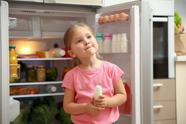 Cute little girl standing with yogurt near open refrigerator at home - Photo, Image